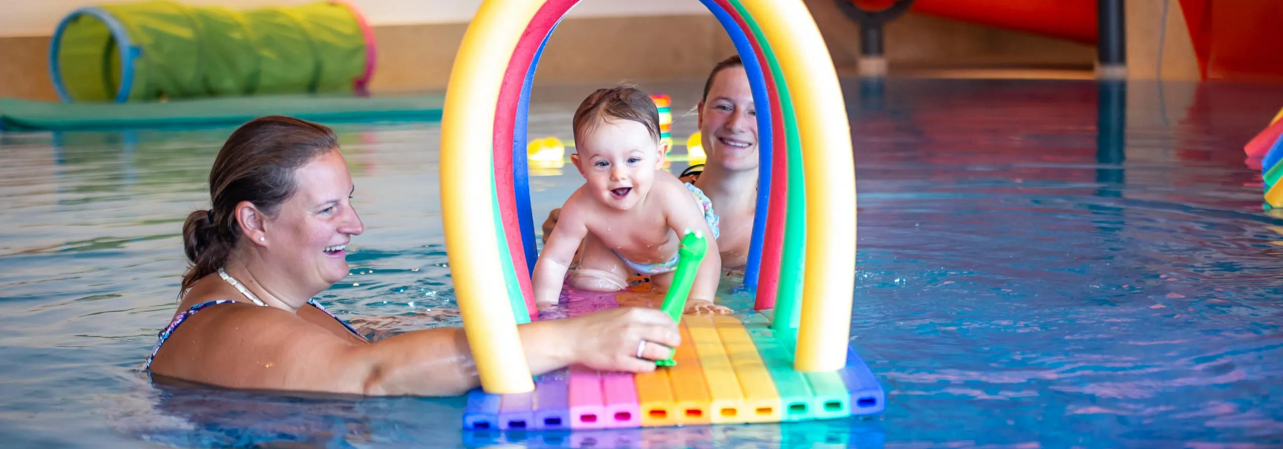 Eltern und Baby spielen im Schwimmbad mit buntem Wasserbogen